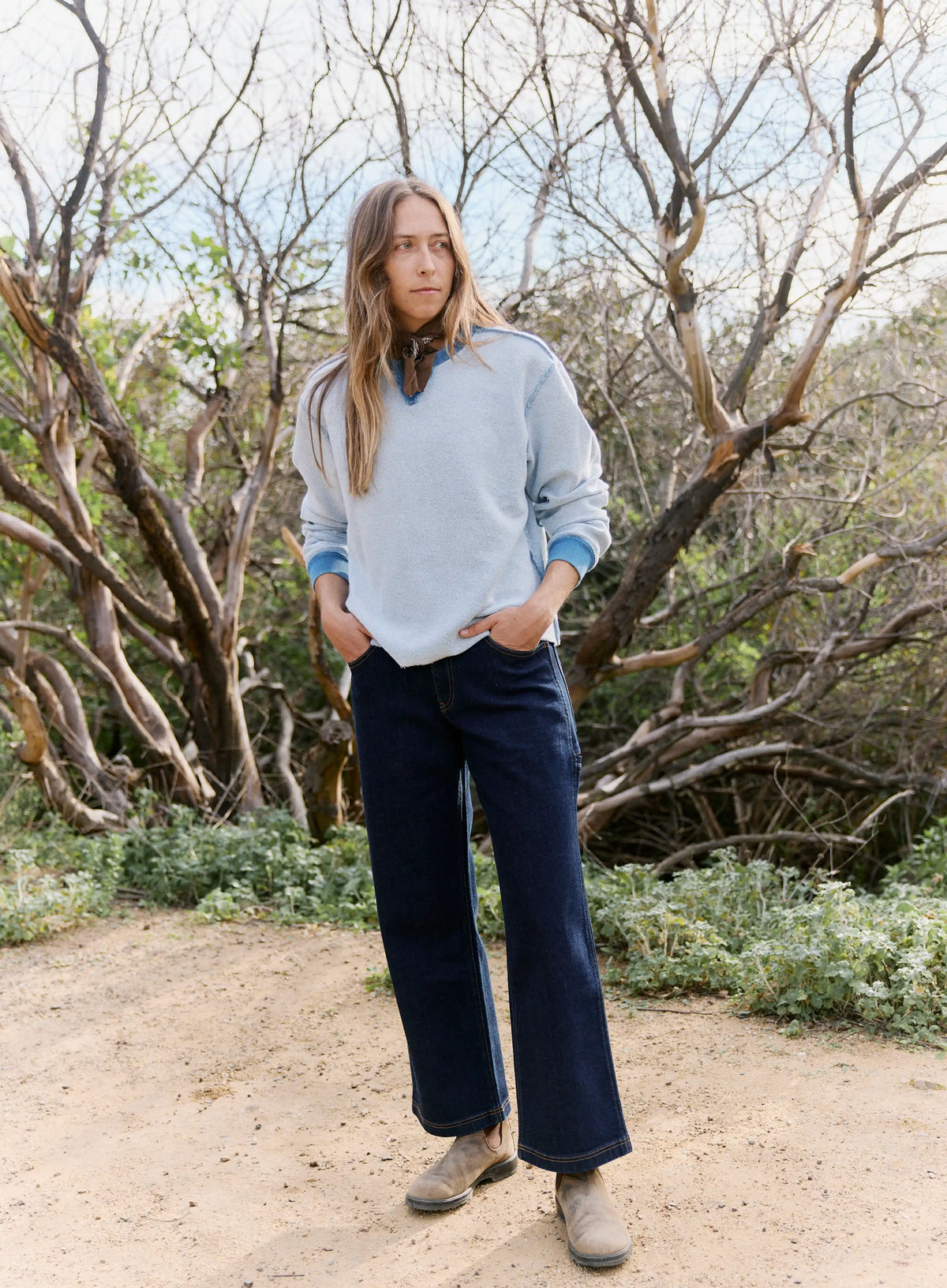 Person wearing a light blue sweater and dark blue pants standing in a natural setting with trees and shrubs.