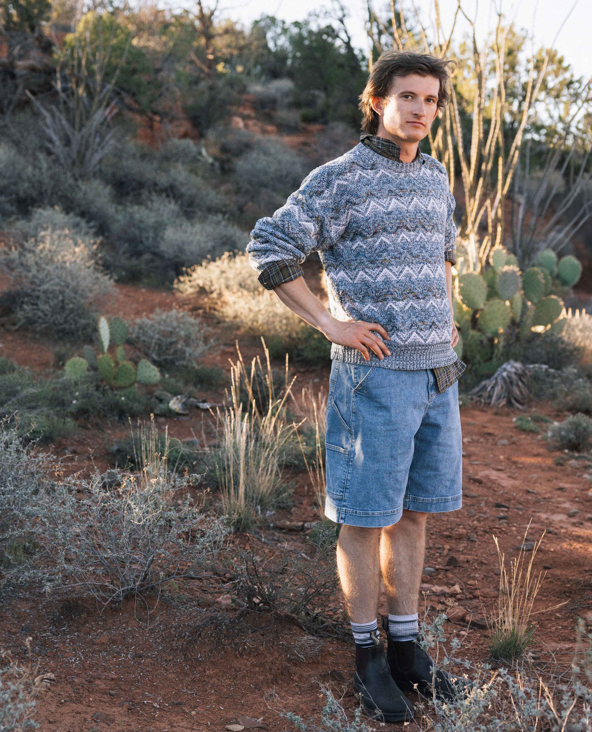 Man wearing a patterned sweater and denim shorts standing in a desert landscape.