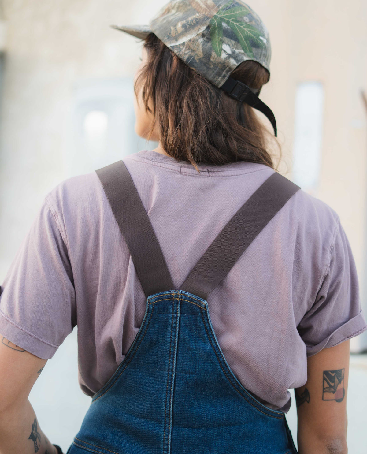 Person wearing a purple shirt and blue overalls with a camouflage cap indoors.