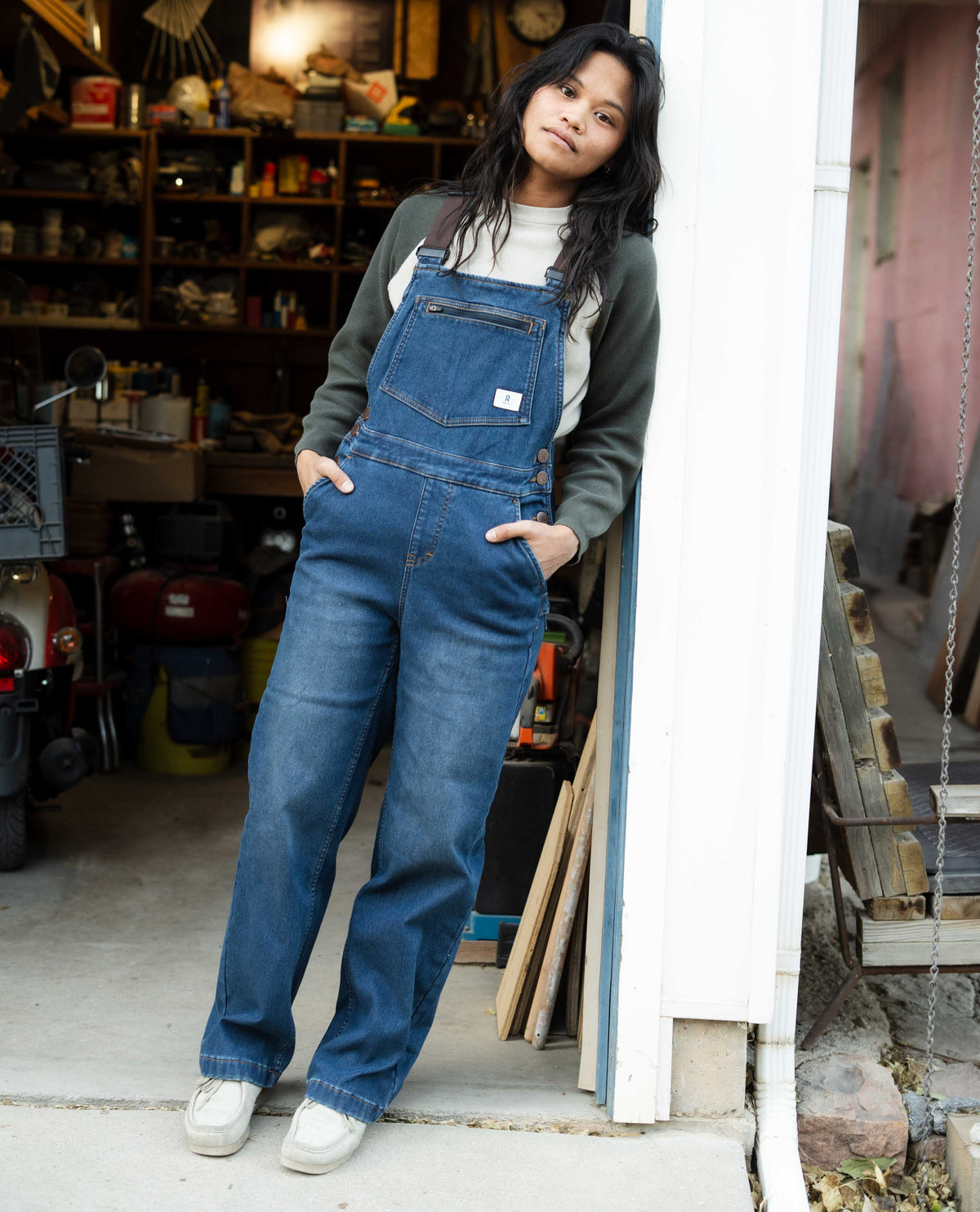 Person wearing blue denim overalls standing in a garage.