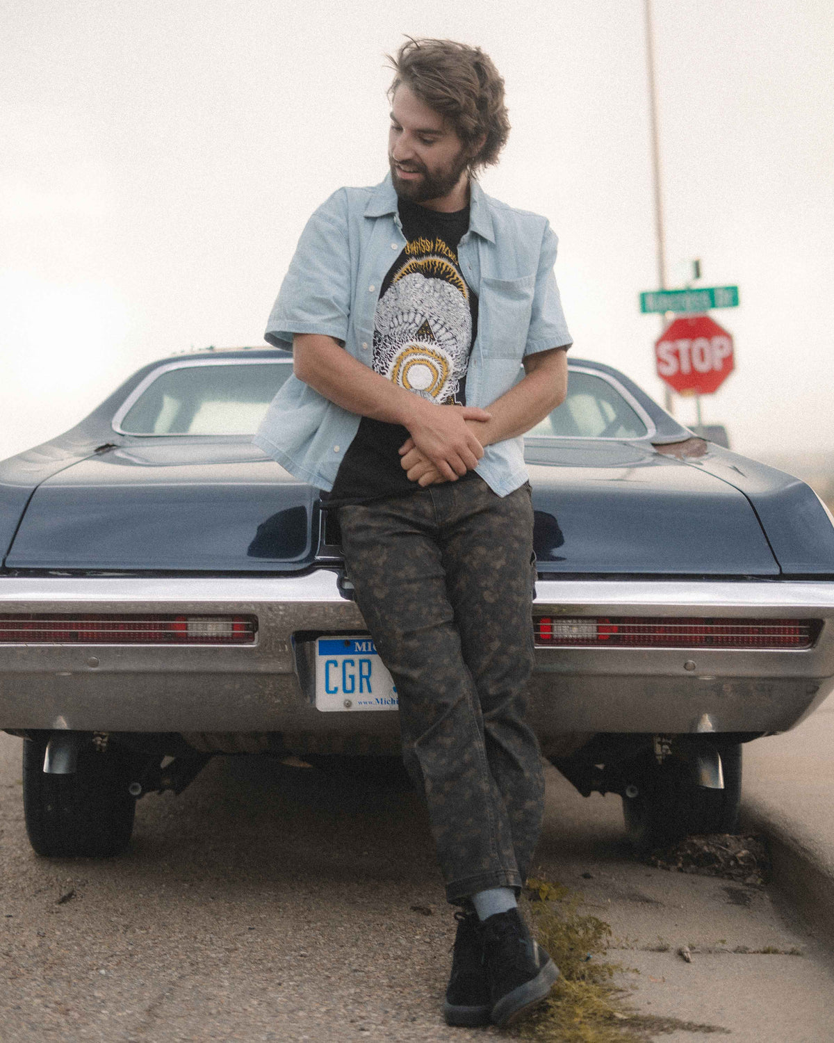 A man leans against the rear of a dark blue classic car outdoors, wearing a light blue button-up shirt over a black graphic t-shirt, dark camouflage pants, and black shoes.