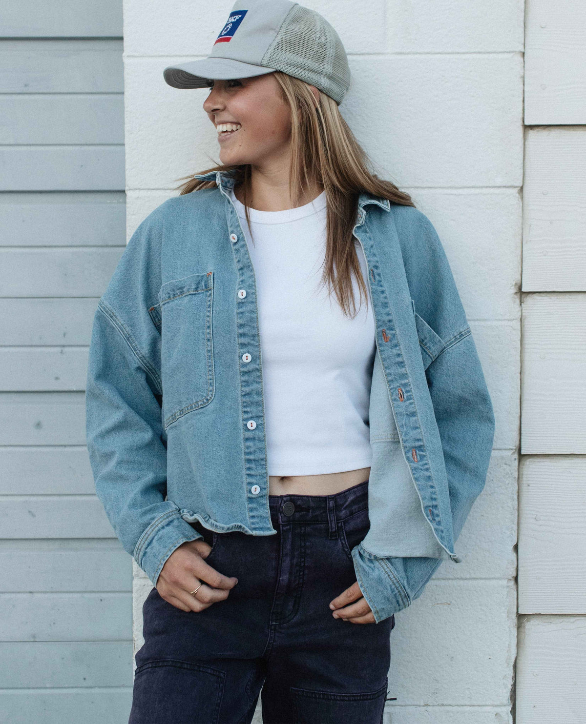 Person wearing a denim jacket, white crop top, and dark pants against a light wooden wall.