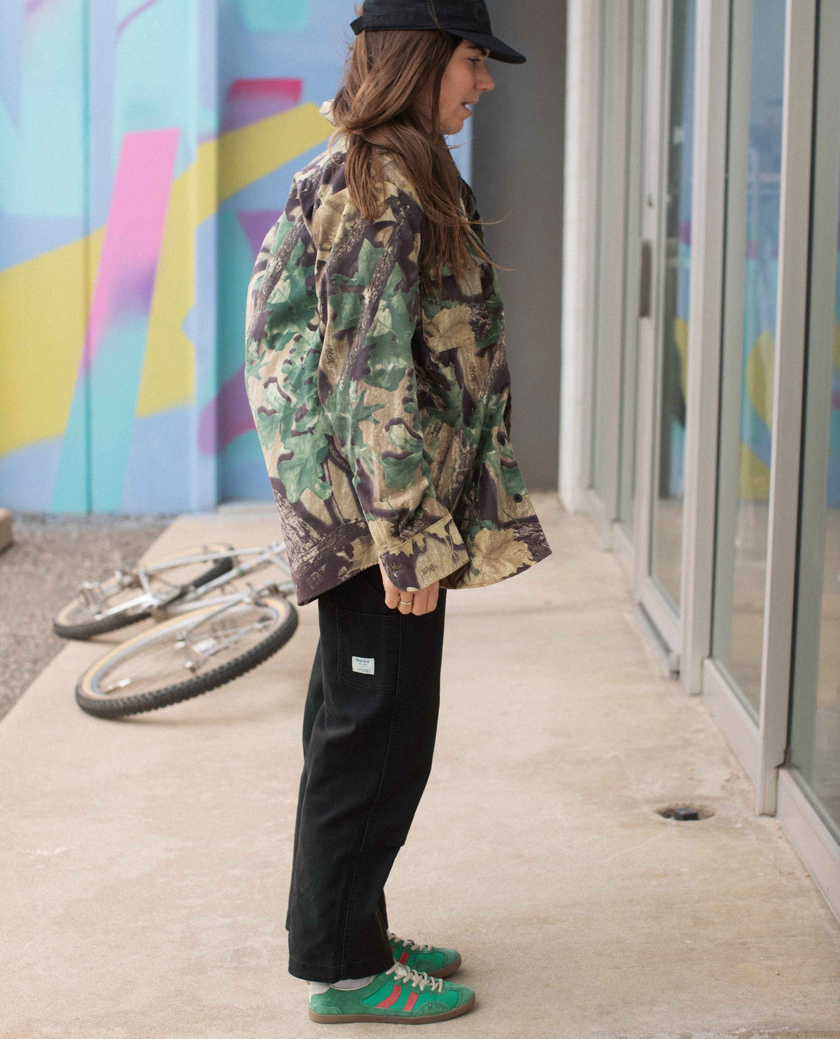 Person wearing a camouflage jacket and black pants standing on a sidewalk with a colorful mural in the background.