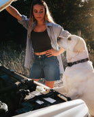 Girl wearing blue denim shorts and black shirt, standing next to dog in front of open hood of a truck.