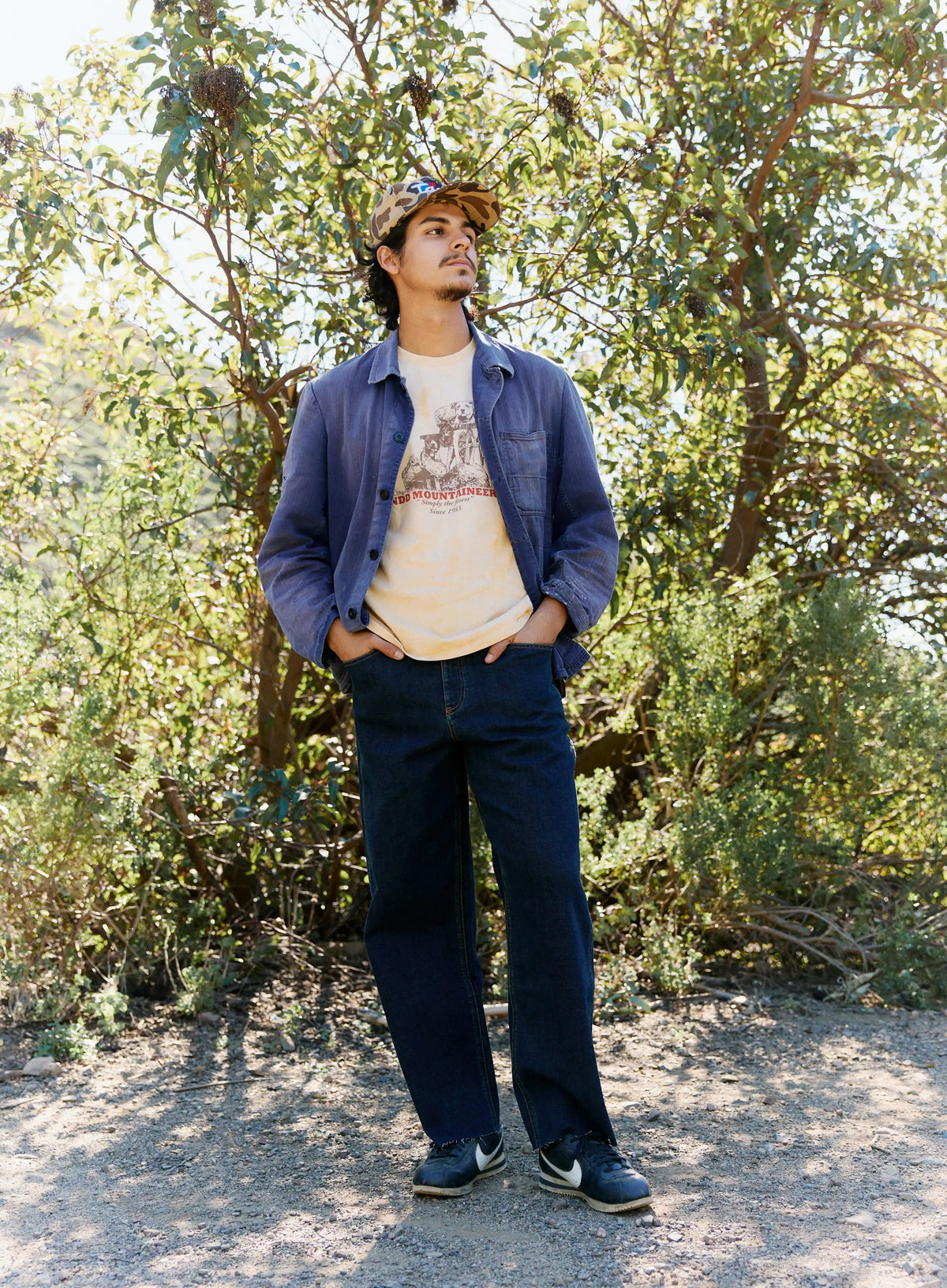 Person wearing a denim jacket and cap standing in a natural setting with trees and shrubs.