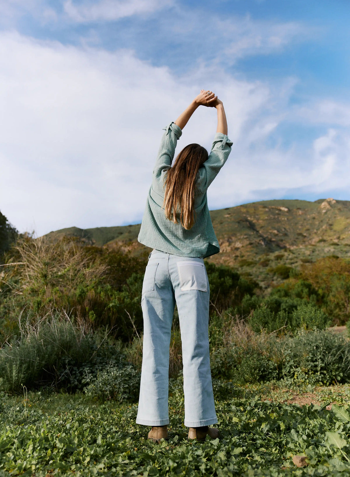 Person standing in a field with arms raised, surrounded by greenery and mountains.
