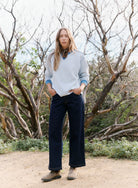 Person wearing a light gray sweater and dark blue jeans standing in a natural setting with trees and shrubs.