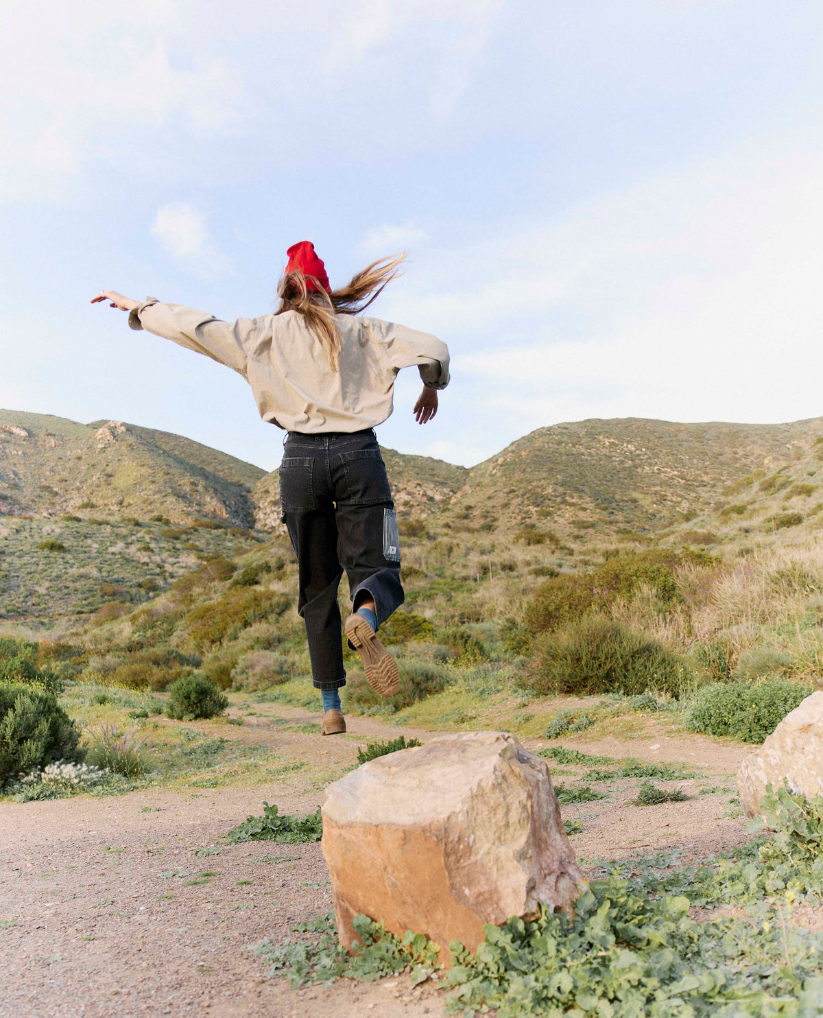 Person in beige jacket and red beanie standing on a rock in a desert landscape