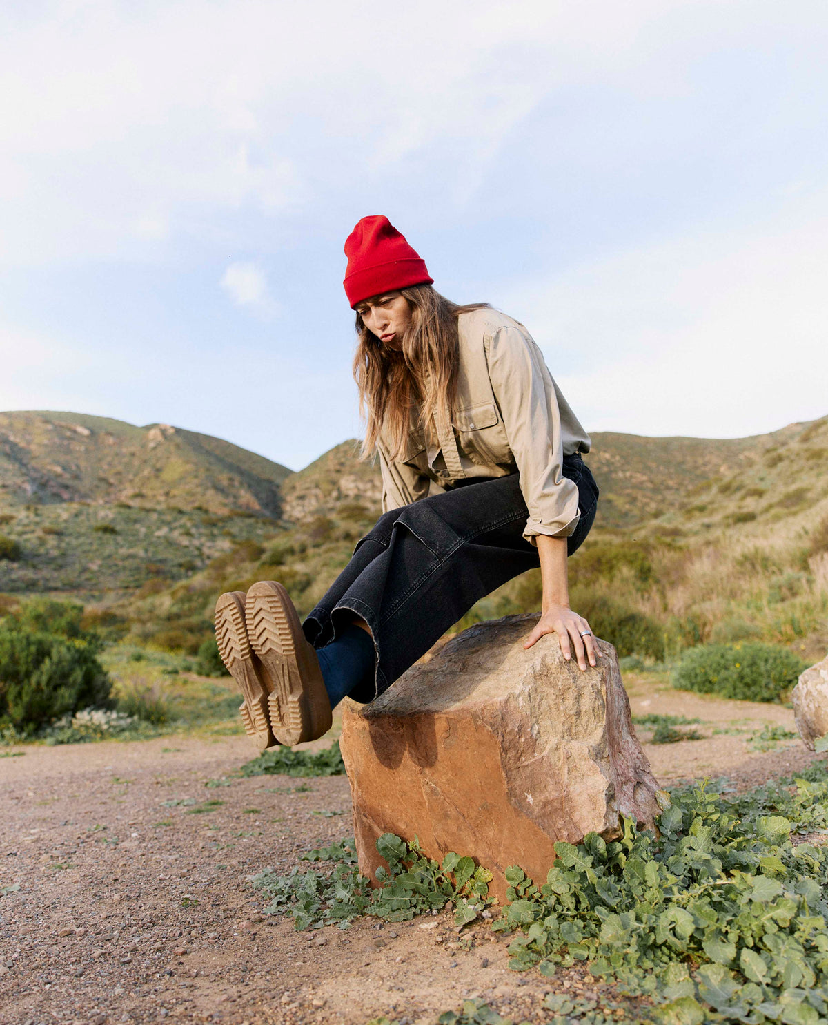 Person wearing a red beanie and beige jacket sitting on a rock in a desert landscape.