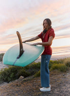 Person holding a teal surfboard on a beach at sunset