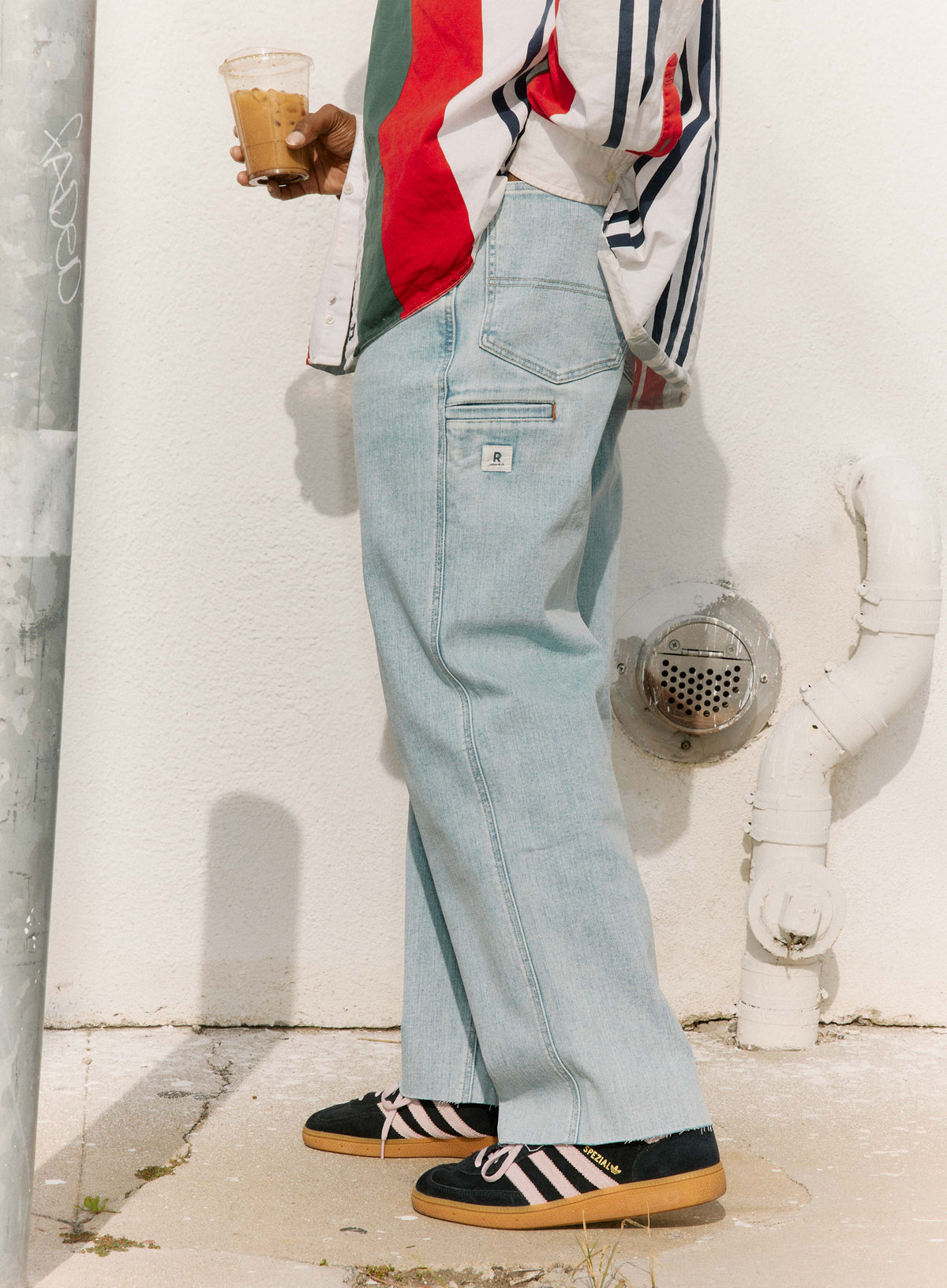 Person wearing light blue jeans and a colorful jacket, standing against a white wall.