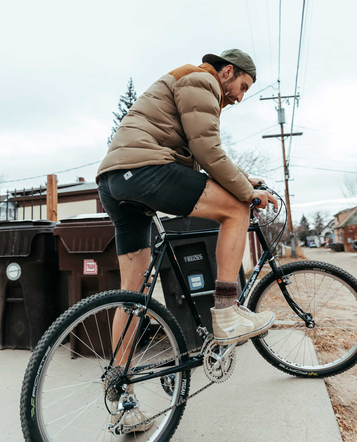 Man outside on bike wearing black shorts and brown jacket in front of trash can