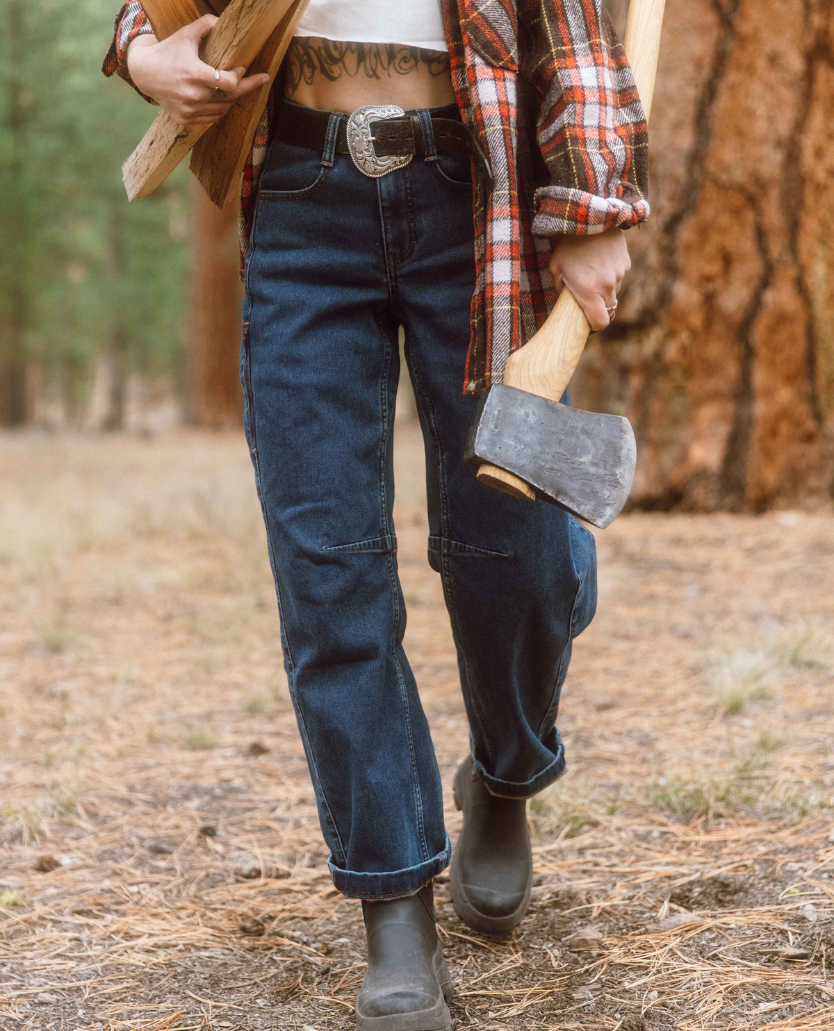 Person in a forest holding an axe, wearing a plaid shirt, jeans, and boots.
