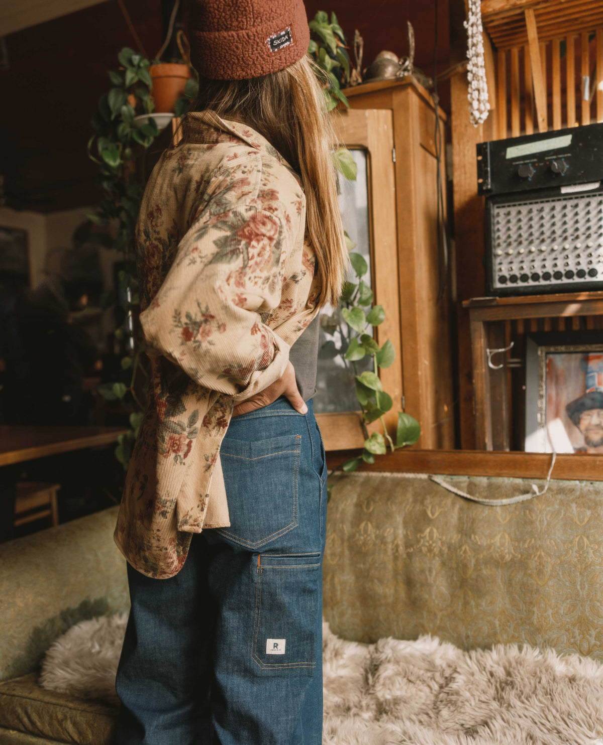 Person wearing a floral shirt and blue jeans in a cozy room with plants and vintage furniture.