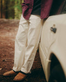Person wearing a maroon shirt and white pants leaning against a vehicle in a forest setting.