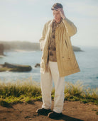 Man standing by a body of water wearing a beige jacket and white pants.