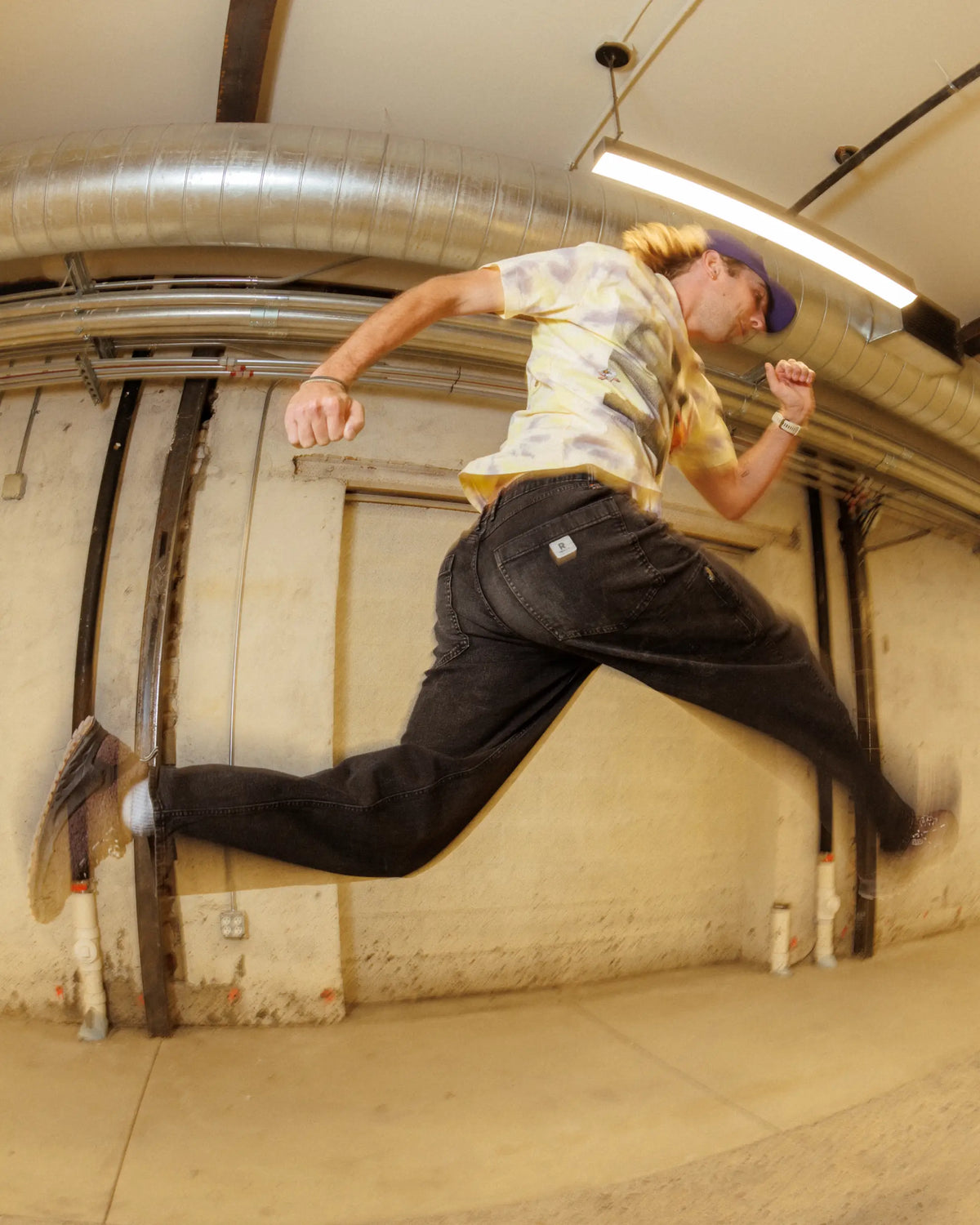 Man jumping in mid-air in front of white wall, wearing blue hat, multi-color shirt, black pants, brown shoes 