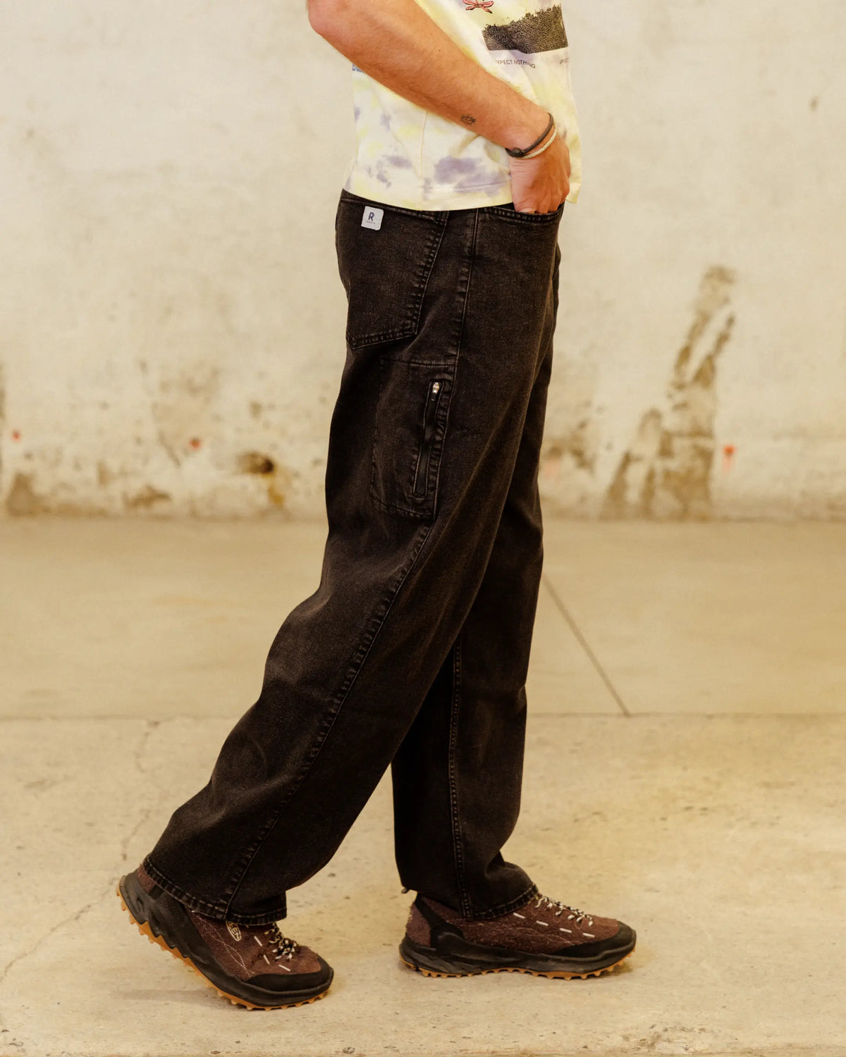 Close up of man's legs wearing black pants, brown shoes, multi-colored shirt, with hands in pockets 