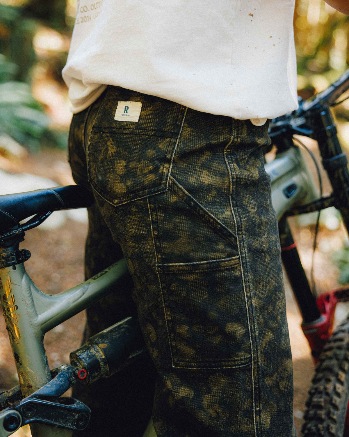 Close-up of a person wearing dark olive camouflage work pants and a white t-shirt, standing next to a mountain bike in an outdoor forest setting.