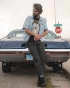 A man leans against the rear of a dark blue classic car outdoors, wearing a light blue button-up shirt over a black graphic t-shirt, dark camouflage pants, and black shoes.