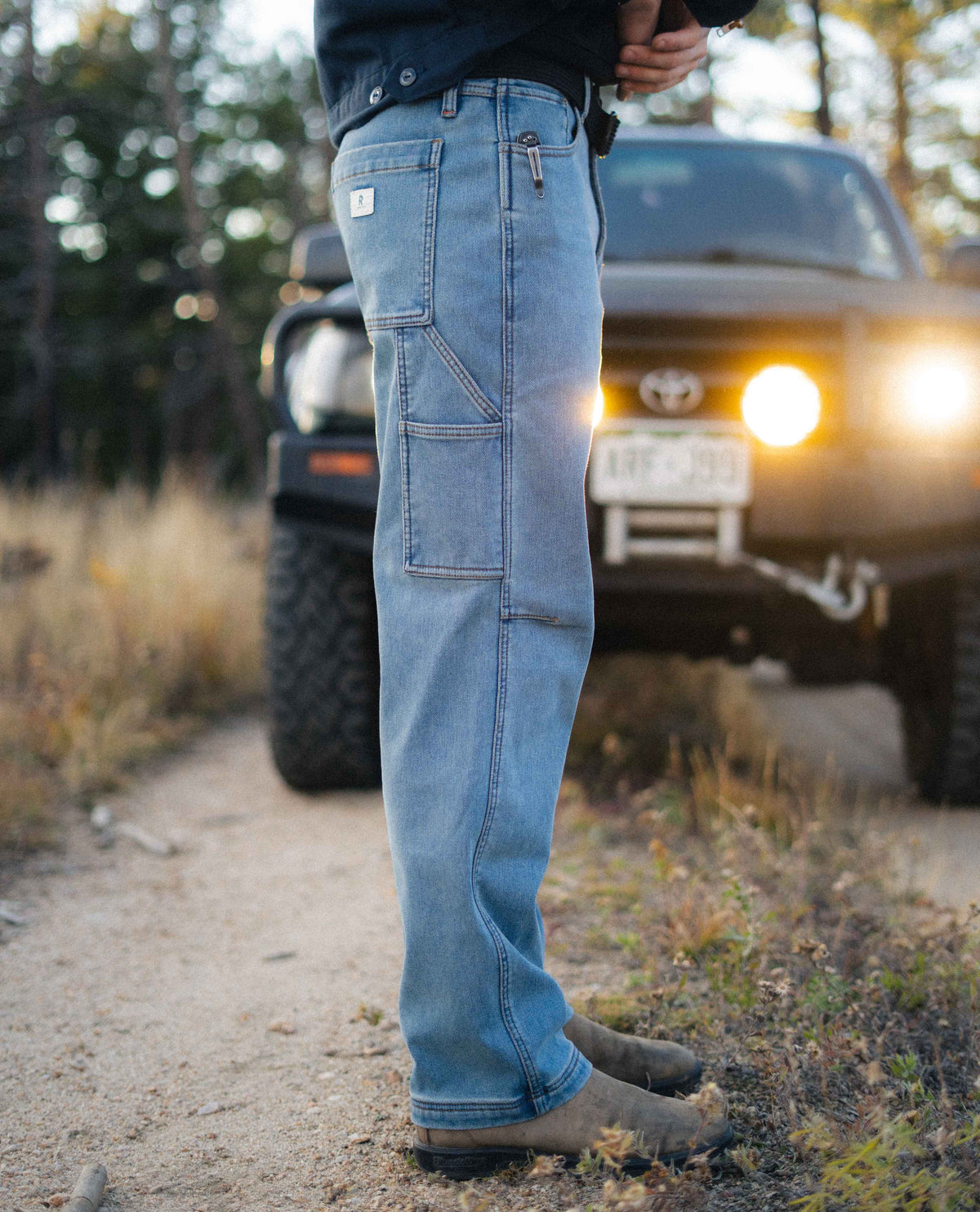 Person wearing blue jeans standing on a dirt road with a vehicle in the background