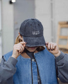 Person wearing a denim cap with 'Ripton' logo, holding it over their face.
