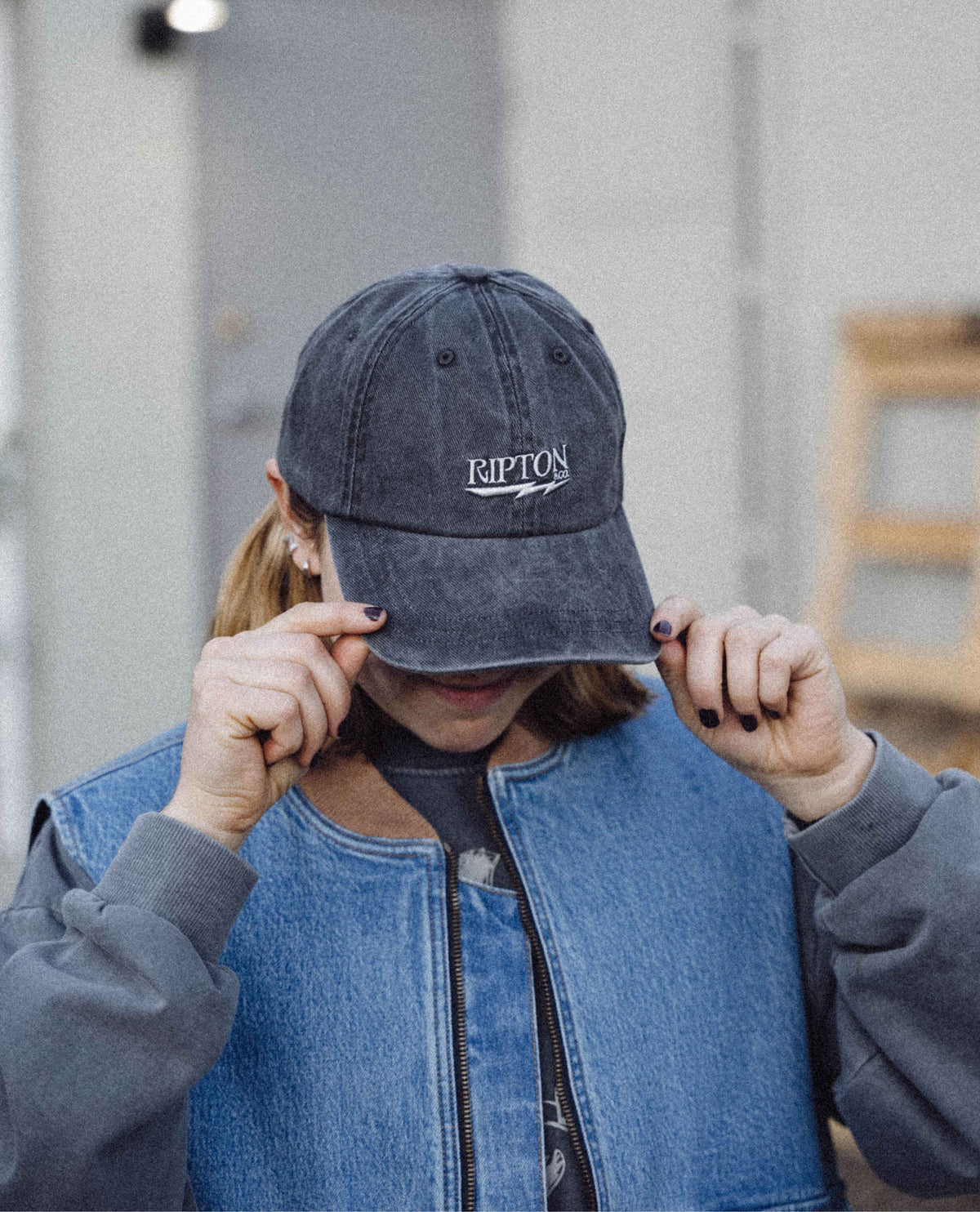 Person wearing a denim cap with 'Ripton' logo, holding it over their face.
