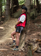 A woman wearing a red vest, white shirt, black shorts, and helmet leaning against a tree