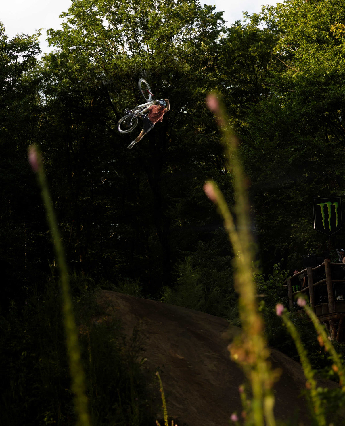 A mountain biker mid-air performing a jump trick in a dense forest, with blurred grass in the foreground and a dirt ramp below.