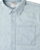 A close-up of a light blue chambray button-down shirt by Ripton & Co, featuring a classic collar, white buttons, and a single chest pocket.