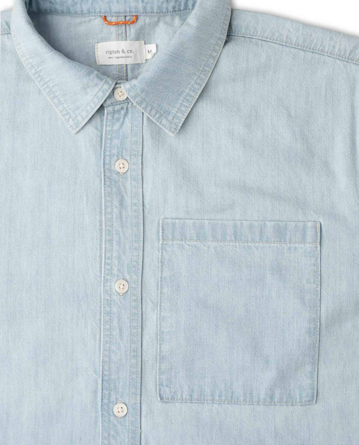 A close-up of a light blue chambray button-down shirt by Ripton & Co, featuring a classic collar, white buttons, and a single chest pocket.