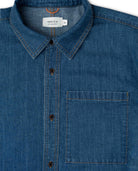 Close-up of a blue denim shirt with a collar and pocket, featuring a brand label.