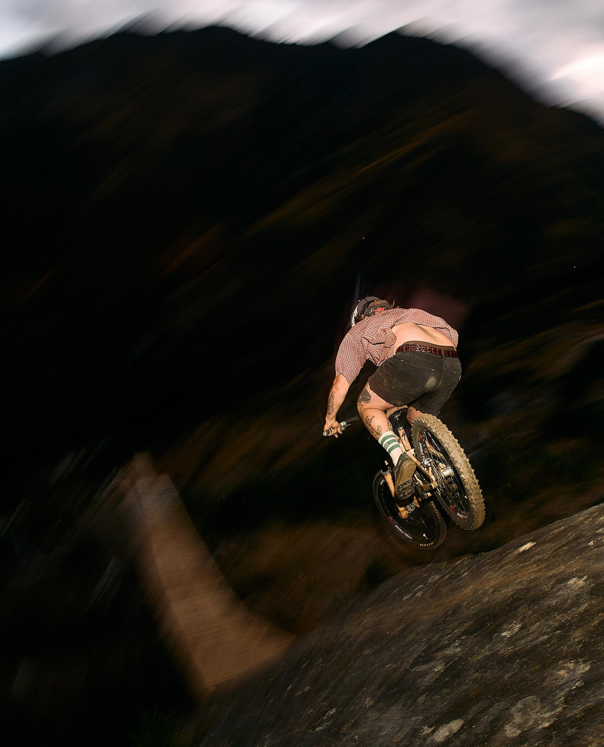 A mountain biker mid-air performing a jump in a dark outdoor setting with motion blur, wearing a pinkish-brown shirt, dark shorts