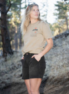 Blonde woman wearing tan shirt, black shorts, with hands in pockets standing outside 