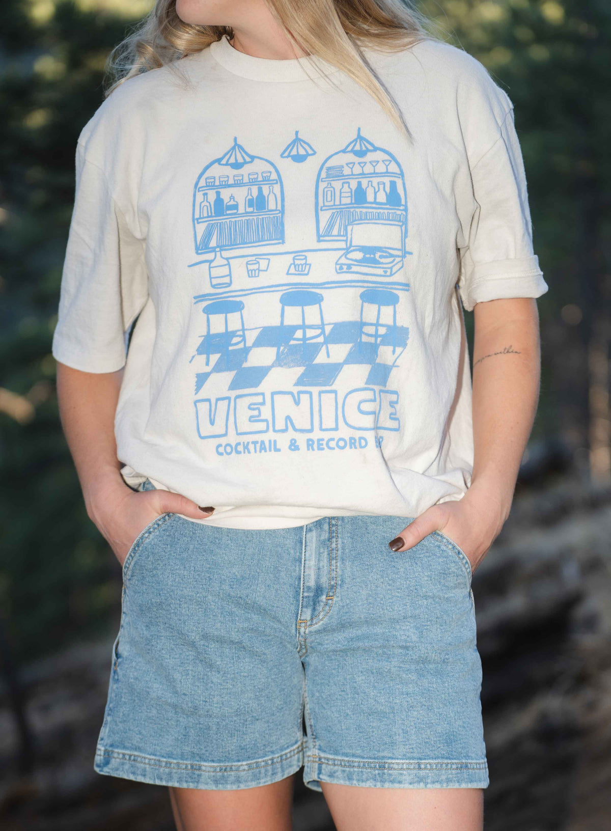 Close up of woman wearing blue shorts and white shirt outside, with hands in pockets 