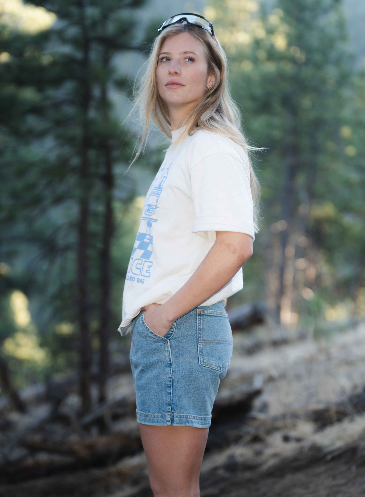 Side view of blonde woman standing outside wearing a white shirt and blue shorts with hands in pockets