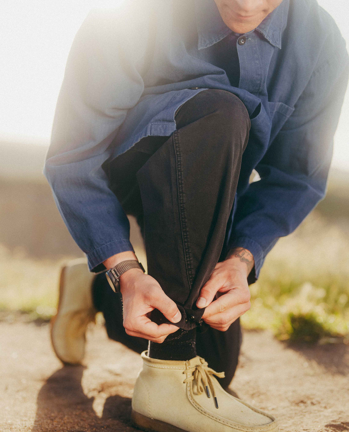 A man outdoors kneeling down to tie the laces of his black pants, wearing a blue button-up shirt and beige boots.