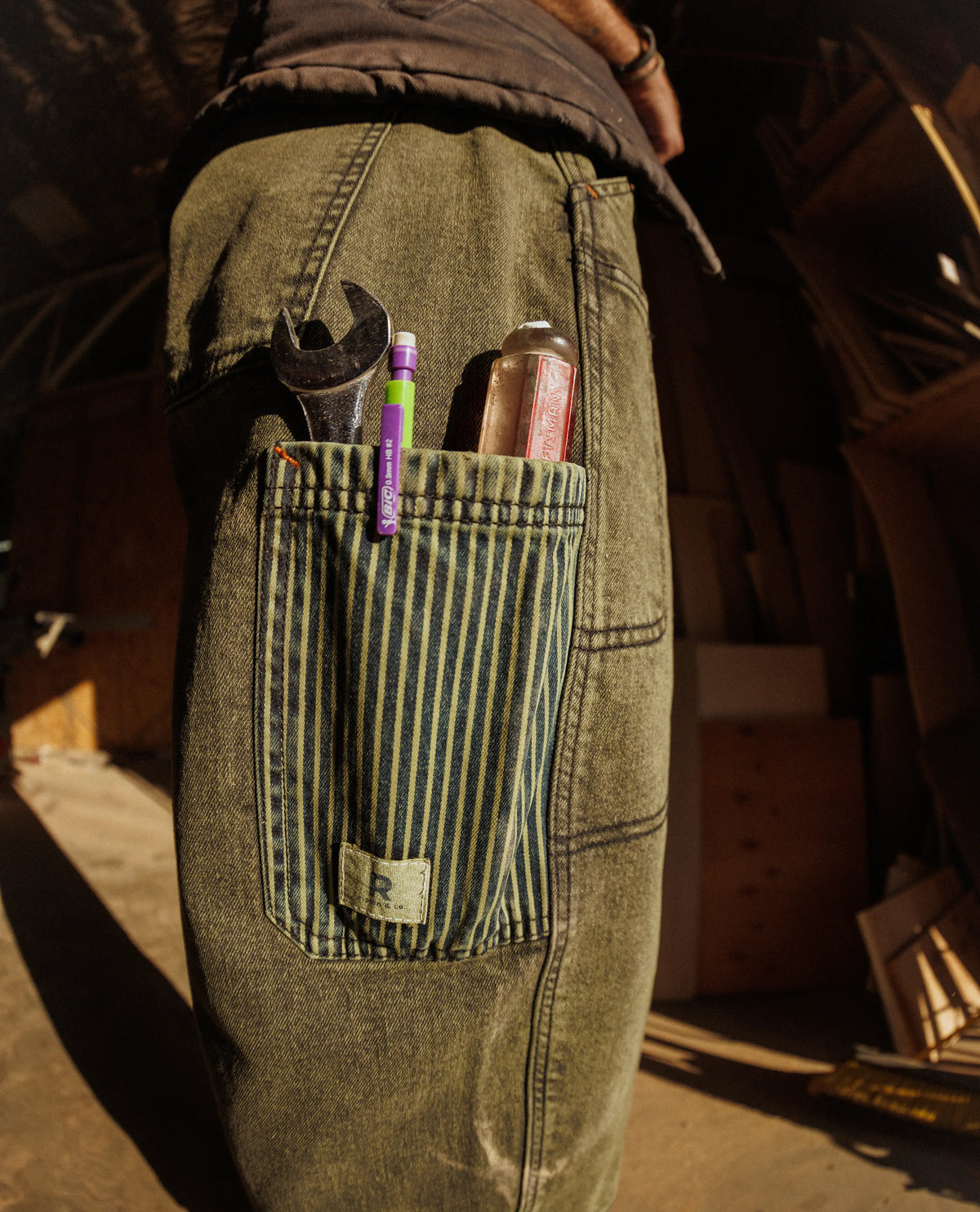 Person wearing green work pants with a pocket containing tools and a can in a workshop setting.