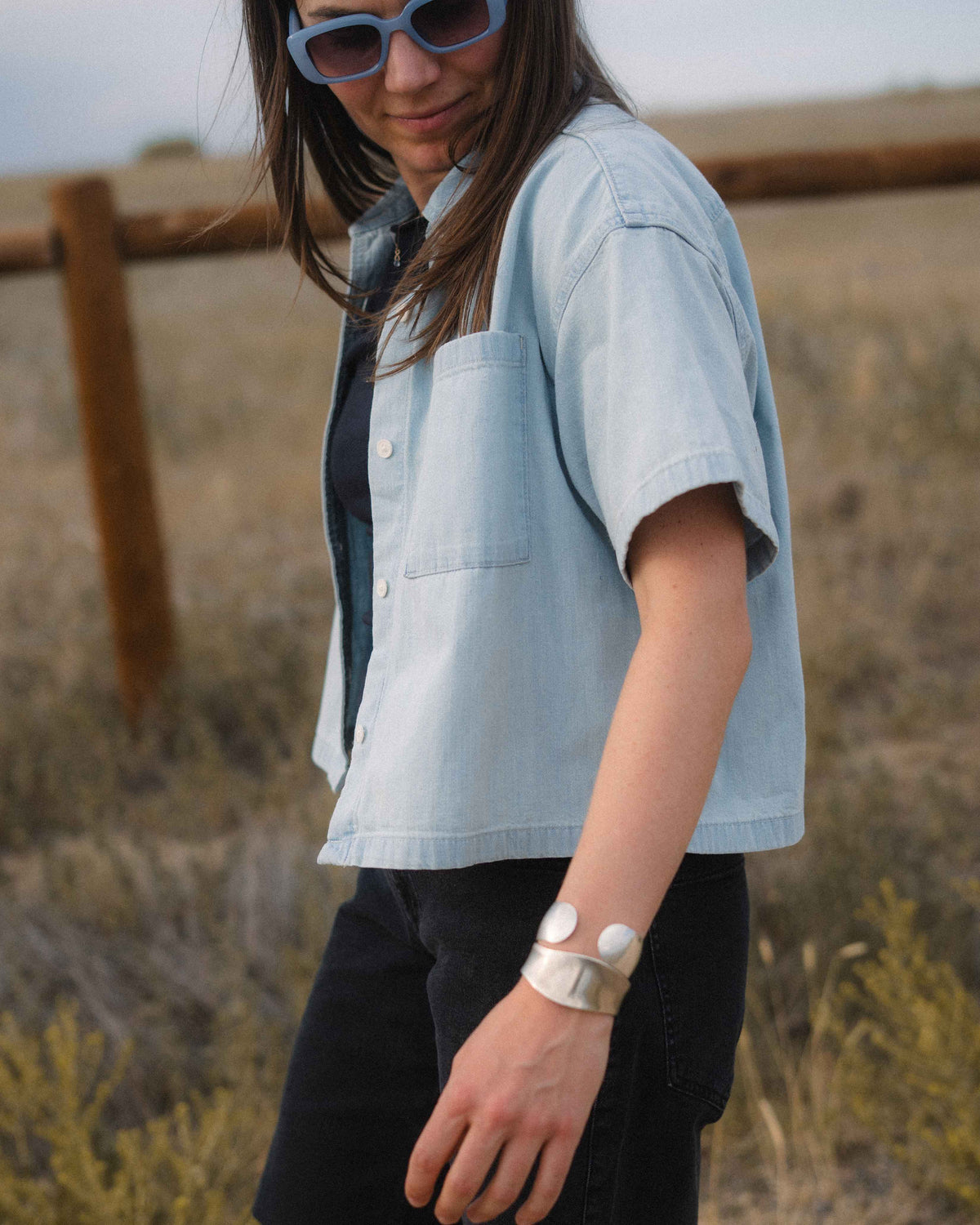 A woman wearing a light blue cropped short-sleeve button-up shirt over a dark top and black pants, standing outdoors in a dry grassy field.