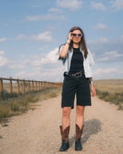A woman stands on a dirt path outdoors wearing a light blue denim shirt, black top and shorts, a black belt with a silver buckle, brown cowboy boots, and sunglasses.