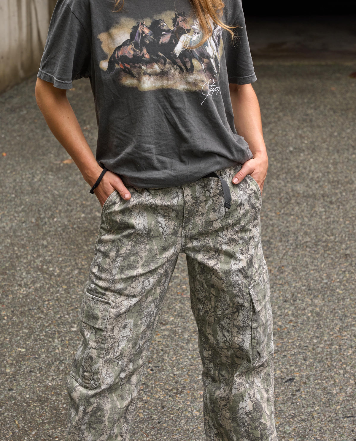 A person wearing a dark gray graphic t-shirt and wide-leg camouflage cargo pants, standing outdoors on a paved surface.