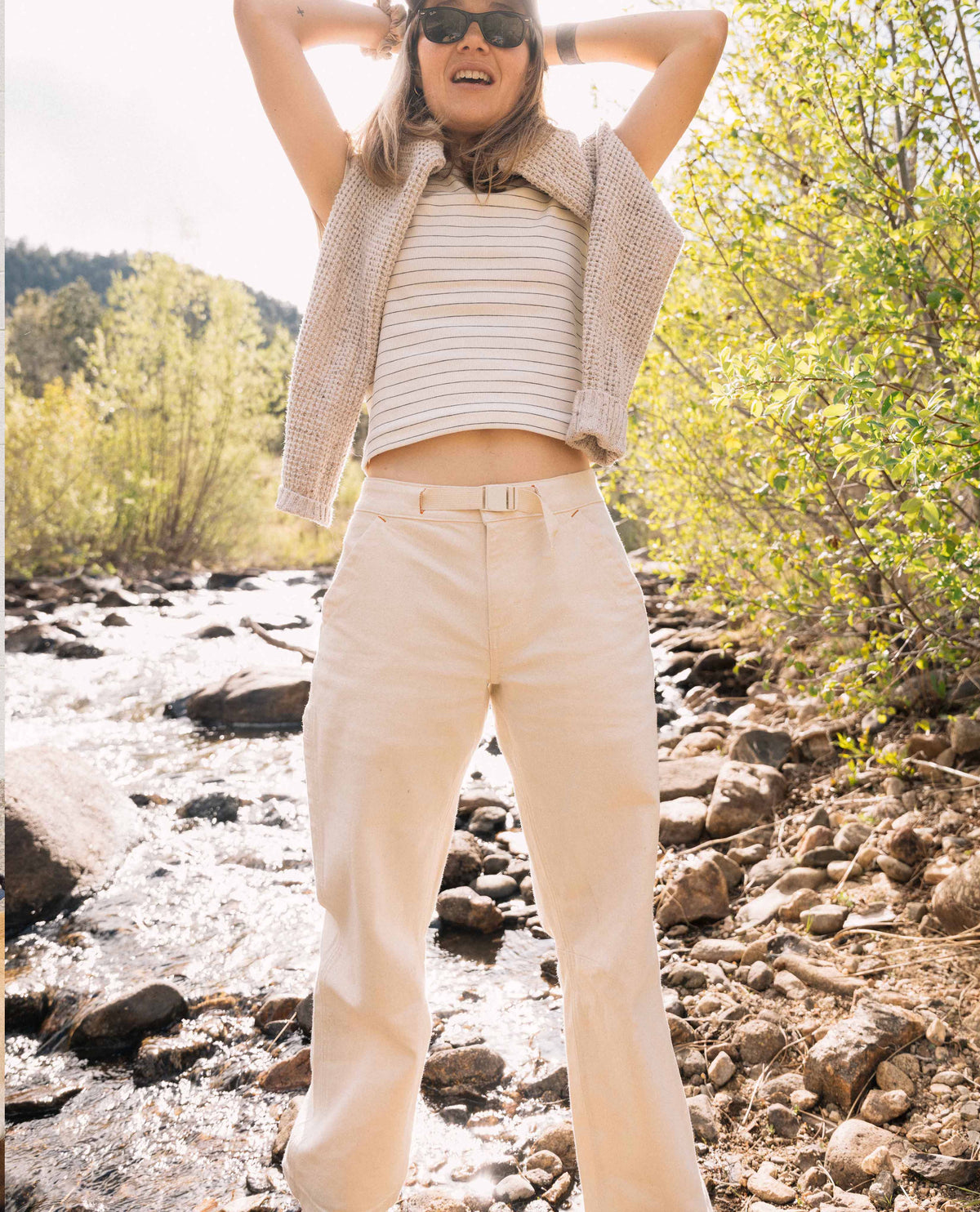 Person standing by a river wearing a striped shirt, vest, and beige pants with sunglasses.
