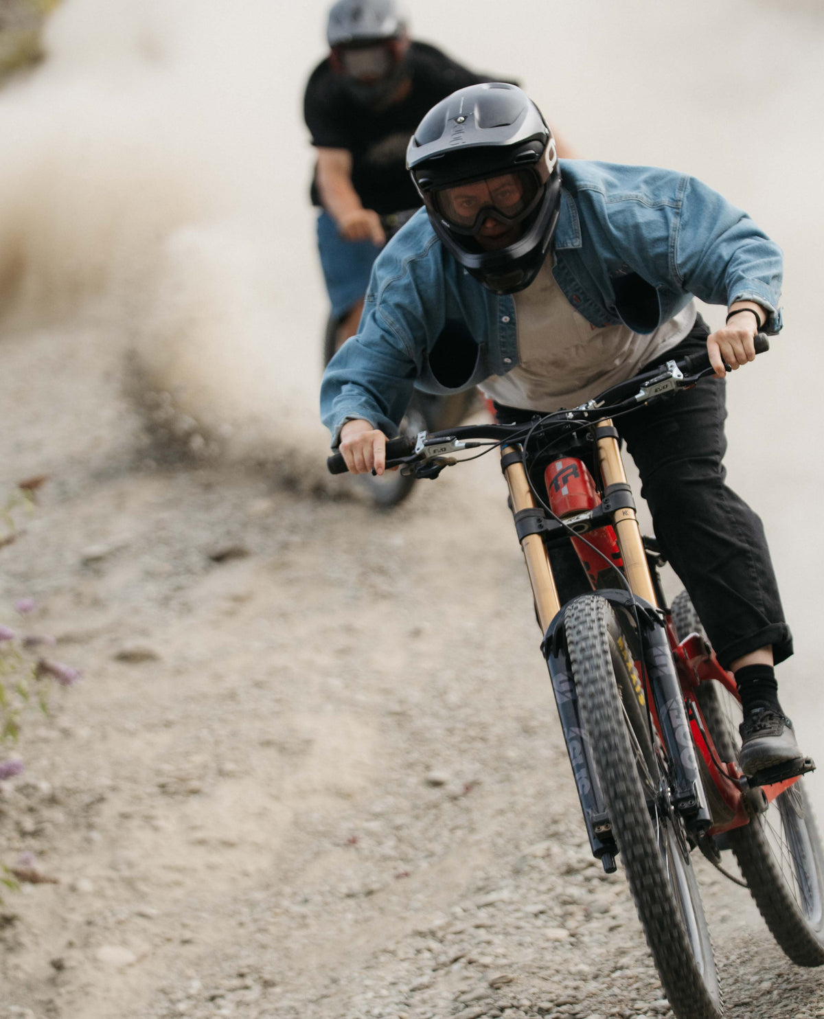 Two people riding bicycles on a dirt path with one person in the foreground wearing a helmet and goggles.