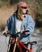 Person wearing a denim jacket and brown cap standing next to a red bicycle outdoors.