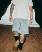 Person wearing light blue shorts and black shoes with tattoos on arms and legs, standing on a sandy surface.
