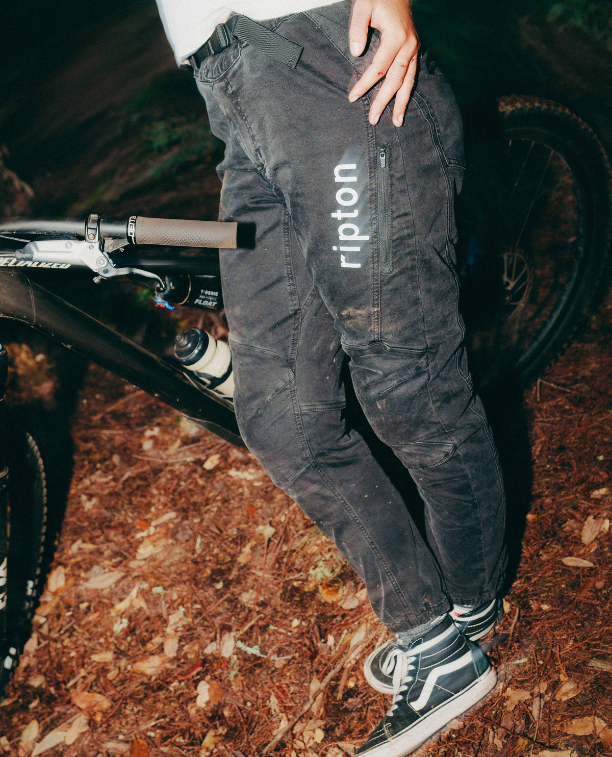 Person wearing black pants with 'ripton' branding, standing next to a bicycle on a trail.