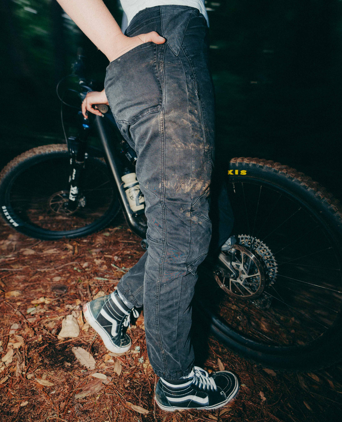 Person wearing dark jeans and sneakers standing next to a bicycle on a forest floor.