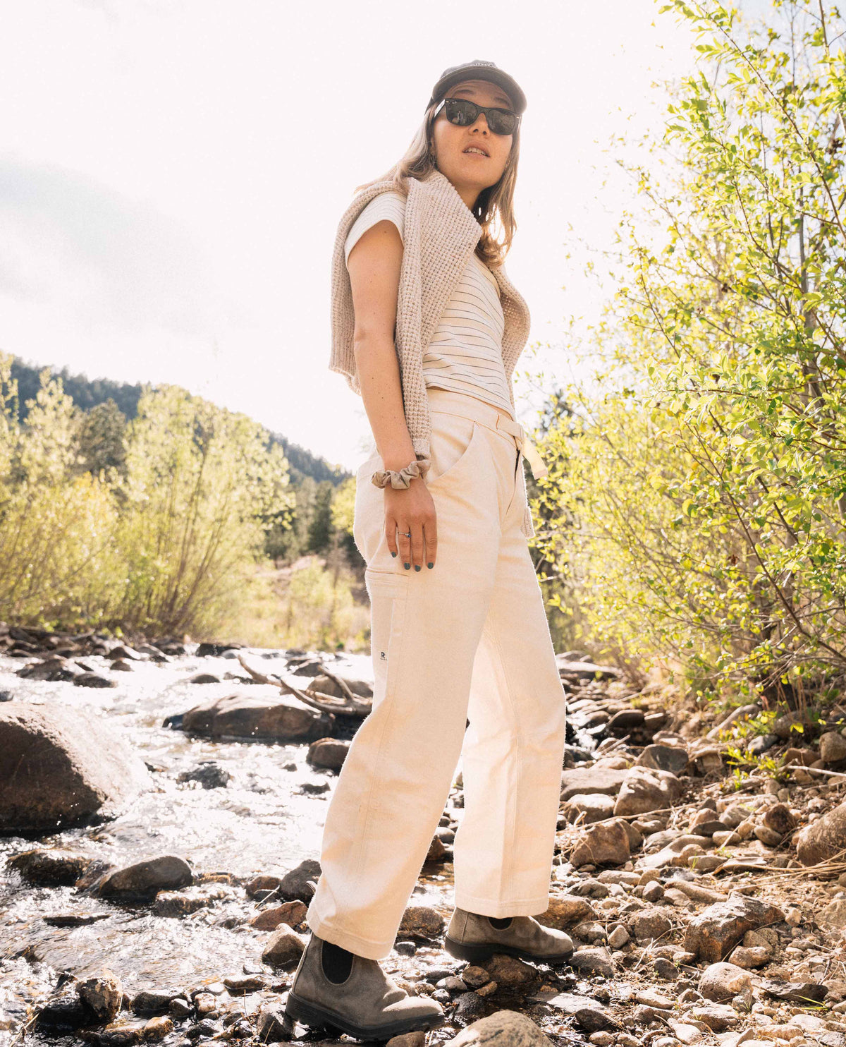 Person wearing a beige outfit standing by a stream with trees in the background