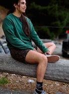 Person sitting on a log outdoors wearing a green sweatshirt and black shorts.