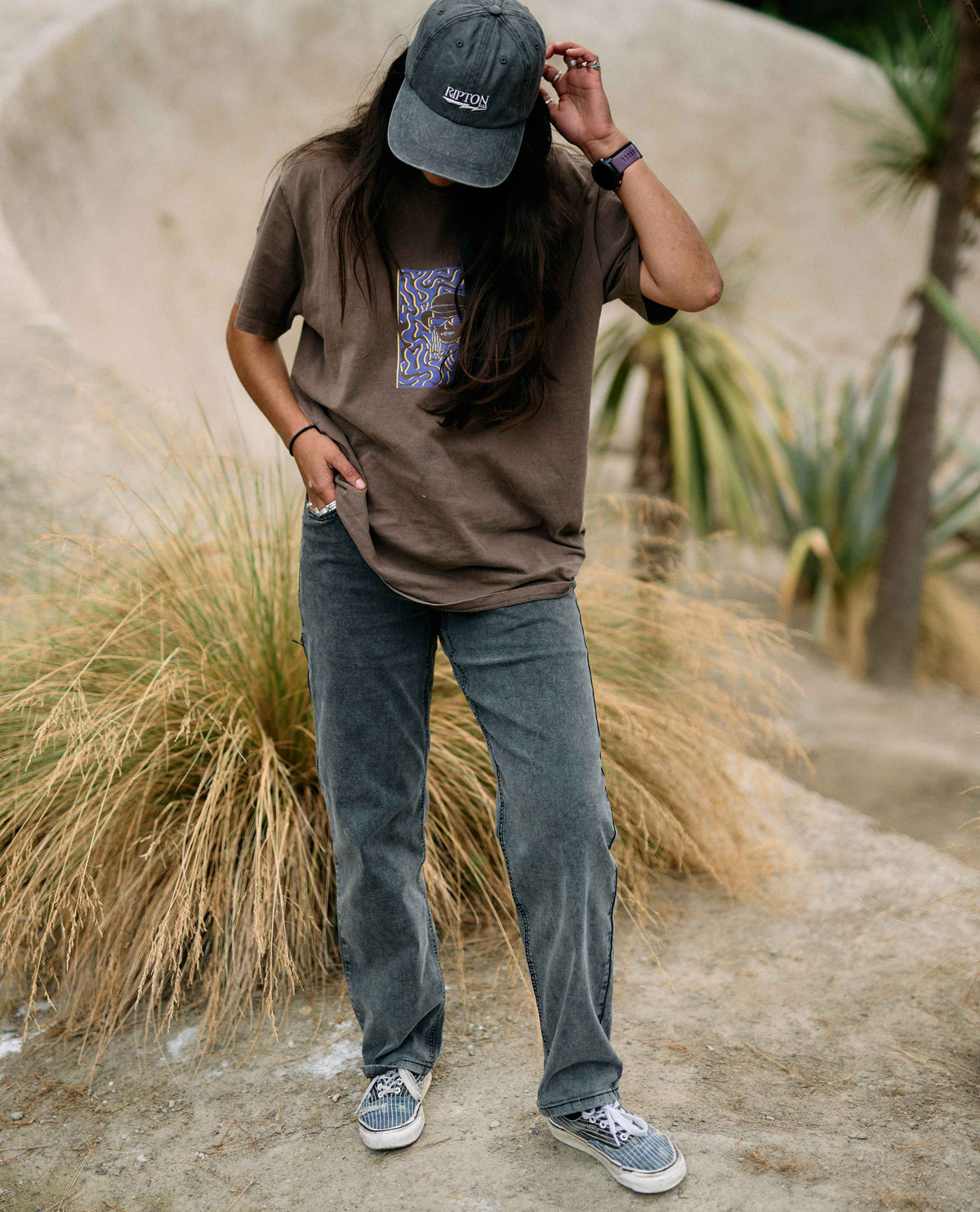 Person wearing a brown t-shirt with a graphic design, blue jeans, and a cap in a natural setting.